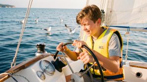 Un adolescent en gilet de sauvetage participe activement à la navigation d’un petit voilier sur une mer calme.